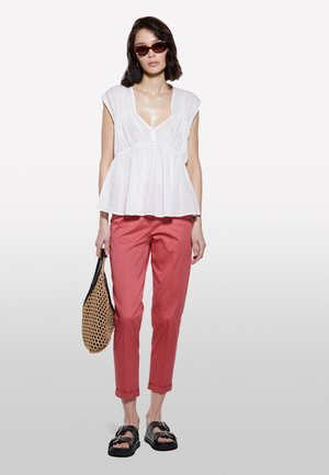 Top bianco a maniche corte con scollatura a V e dettagli arricciati, abbinato a pantaloni corallo. Gli accessori includono sandali neri e una borsa intrecciata.