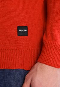 Gros plan sur une personne portant un pull en maille rouge avec une étiquette noire "ONLY & SONS" au-dessus d'un jean bleu foncé.
