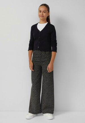 Jong meisje staat, gekleed in een zwarte knooptrui, een witte shirt, een wijde leopardprint broek en witte sneakers tegen een effen achtergrond.