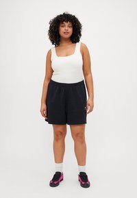 Juodos šortai su elastinga juosmeniu, derinami su balta背tank topu. Modelis dėvi juodus ir rožinius sportinius batus bei baltas kojines.