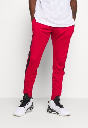 Mężczyzna ubrany w czerwone wąskie spodnie dresowe z czarnymi bocznymi paskami oraz białe wysokie trampki z czarnymi sznurowadłami, stojący z rękami w kieszeniach.