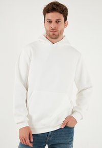 Hvid hættetrøje lavet af blødt stof med en frontlomme og ribstrikkede manchetter, kombineret med blå jeans.