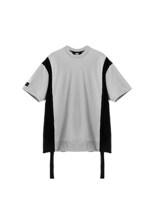 Joven con rastas que lleva una camiseta gris extragrande con paneles laterales negros, pantalones anchos negros con detalle de cremallera y zapatos negros texturizados.