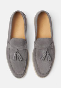 Mocassini in suede grigio con design a mocassino, caratterizzati da una cucitura rialzata e due nappine decorative sul davanti. Suola in gomma leggera.