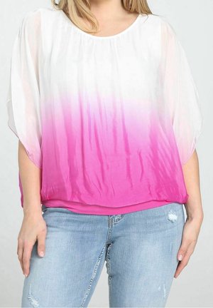 Femme portant un haut blanc ample avec un dégradé rose et un jean déchiré bleu clair, debout devant un fond clair uni.