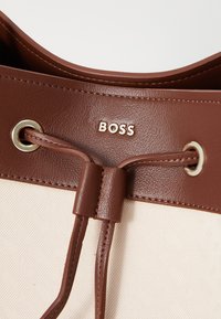Chiusura a coulisse in pelle marrone con accessori dorati. Tessuto di colore crema dalla texture con un motivo sottile. Nome del marchio "BOSS" impresso.