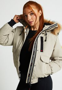 Bēža pūkveida jaka ar kažokādas odere kapuci, melnā krāsā akcenti, rāvējslēdzējs ar logotipu un priekšējā kabata. Gluda tekstūra, ikdienišķs dizains.