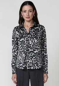 Una donna indossa una blusa a stampa leopardata bianca e nera con colletto, abbinata a pantaloni scuri, in piedi su uno sfondo neutro.