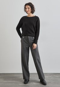Schwarzer gestrickter Pullover mit weiter Ausschnittkante, kombiniert mit hochtaillierten grauen Hosen mit Struktur, die über Knopfdetails und weite Beine verfügen. Schwarze Schuhe.