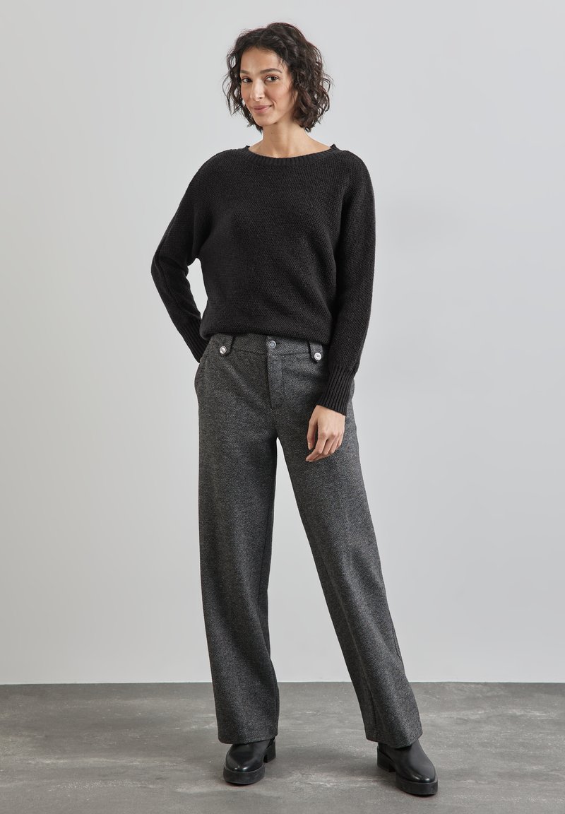 Schwarzer gestrickter Pullover mit weiter Ausschnittkante, kombiniert mit hochtaillierten grauen Hosen mit Struktur, die über Knopfdetails und weite Beine verfügen. Schwarze Schuhe.