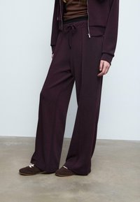 Burgunderfarbene weit geschnittene Hose aus weichem Stoff mit Kordelzugbund. Kombiniert mit einem dunkelbraunen Oberteil und braunen Slippers. Glatte Textur.