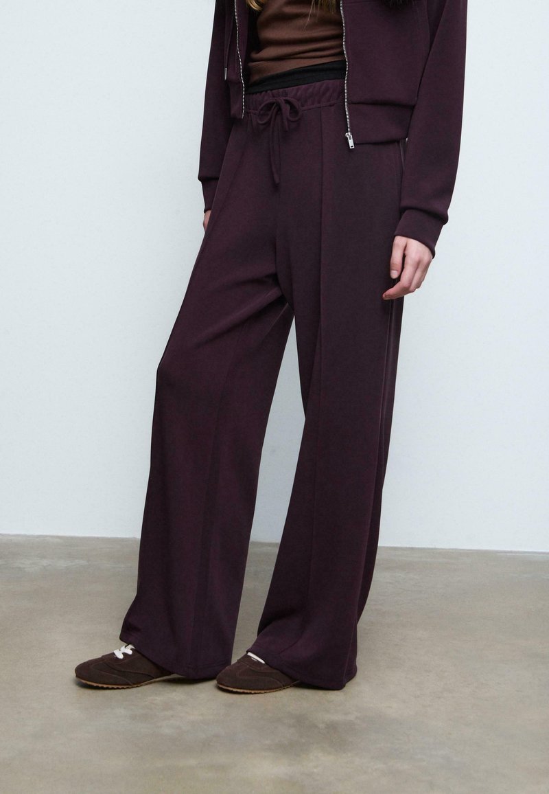 Burgunderfarbene weit geschnittene Hose aus weichem Stoff mit Kordelzugbund. Kombiniert mit einem dunkelbraunen Oberteil und braunen Slippers. Glatte Textur.