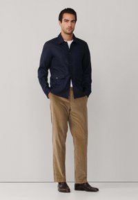 Marineblauwe knopenjack met twee voorkruiszakken, gecombineerd met lichtbruine corduroy-broeken en donkerbruine schoenen, staande tegen een neutrale achtergrond.