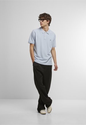 Ung mann iført lys blå polo, svarte vide bukser, briller og hvite joggesko, står med hendene i lommene mot en ensfarget bakgrunn.