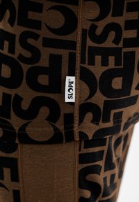 Braune Leggings mit einem großen schwarzen Textmuster und einem weißen "SLOPE"-Logoetikett. Sie sind mit einer glatten Textur und enger Passform gestaltet.
