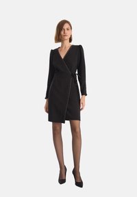 Czarna sukienka kopertowa z długimi rękawami, zdobiona złotymi ćwiekami wzdłuż dekoltu i przodu, z paskiem w talii do związania.