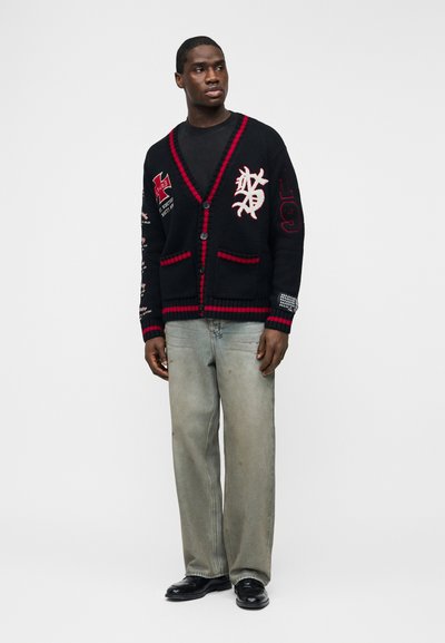 Uomo che indossa un cardigan nero con rifiniture rosse e toppe, una maglietta nera, jeans grigi slavati e scarpe nere, in piedi contro uno sfondo semplice.