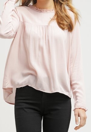 Vrouw draagt een lichtroze blouse met lange mouwen, geruchte kraag en manchetten, gecombineerd met een zwarte broek, tegen een effen achtergrond.