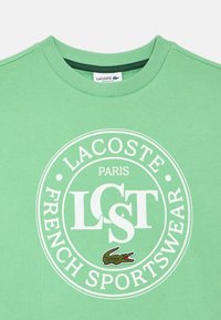 Zelená mikina s ribovým výstrihom, s bielym kruhovým logom s nápisom „LACOSTE PARIS FRENCH SPORTSWEAR“ a vyšívaným detailom krokodíla.