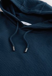 Laivastonsininen huppari, jossa on ribbineulosta, säädettävä kiristysnaru ja metalliset nyörinpäälliset. Sisäpuolella huppua näkyvissä oleva merkki.