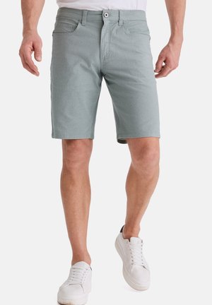 Mann trägt hellgraue knielange Shorts und weiße Sneaker und steht vor weißem Hintergrund.