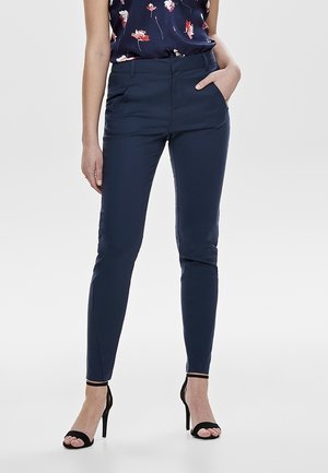 Femme portant un pantalon slim bleu marine, un haut sans manches à motif floral et des sandales à talons hauts noires, une main dans la poche, debout sur un fond blanc.