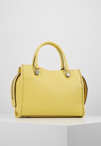 Keltainen nahkainen käsilaukku, jossa on pyöreät kaksoiskahvat ja hopeiset yksityiskohdat, esillä valkoisella jalustalla harmaata taustaa vasten.