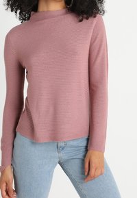 Top rosa a maniche lunghe con texture a coste e collo alto, abbinato a jeans azzurri. Tessuto morbido e vestibilità comoda.