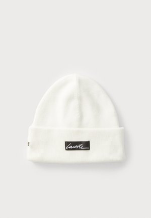 White ribbed knit beanie with folded cuff featuring a black label with white "Lacoste" text on front center.