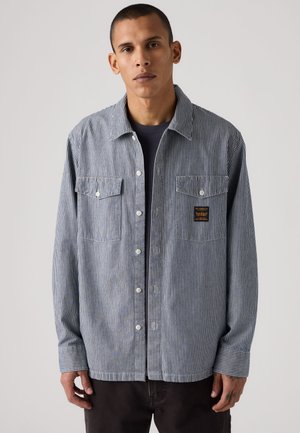 Jeune homme aux cheveux courts portant une chemise rayée bleue et blanche boutonnée sur un t-shirt foncé, debout devant un fond clair uni.