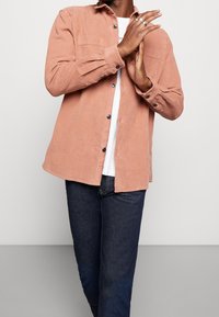 Man med en ljusbrun manchesterknapp-up skjorta över en vit t-shirt och mörkblå jeans, med händerna knäppta vid bröstet.