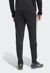 Μαύρο αθλητικό παντελόνι με λευκές λωρίδες στο πλάι, στενή γραμμή και ελαστική μέση. Φορεμένο με μαύρα και πράσινα ποδοσφαιρικά παπούτσια.