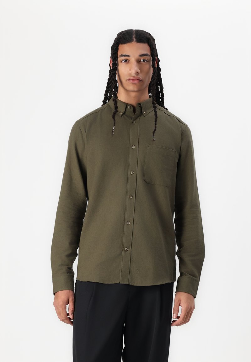 Camisa de botones verde oliva en tela texturizada, con bolsillo en el pecho, mangas largas y dobladillo curvo. Diseño minimalista con acentos sutiles.