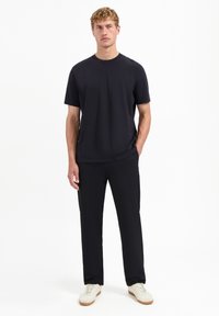 Zwarte crew-neck t-shirt met korte mouwen, gecombineerd met zwarte broek. Het model draagt witte sneakers met lichtbruine zolen.