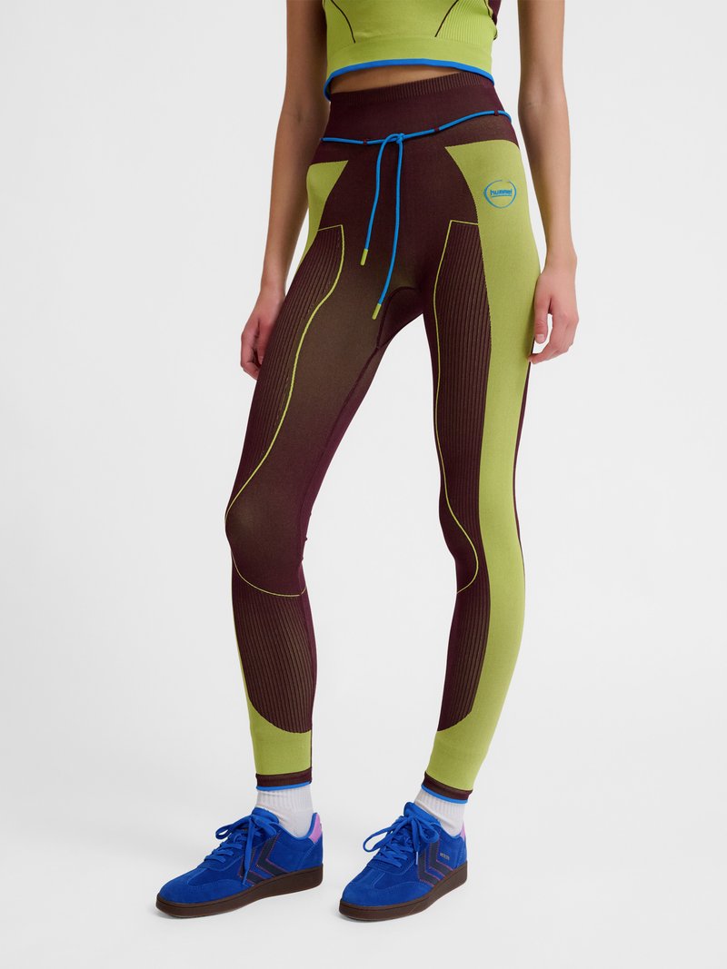 Person wearing green and brown color-blocked leggings with blue drawstring, white socks, and blue sneakers, standing against plain background.