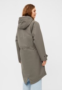 Cappotto grigio di lunghezza media con cappuccio, vita aderente, caratterizzato da uno spacco posteriore e polsini con bottoni, realizzato in un tessuto liscio e resistente all'acqua.