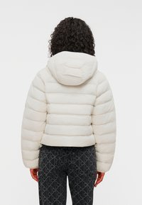 Λευκό, φουσκωτό μπουφάν με κουκούλα και ελαστικές άκρες; συνδυασμένο με σκούρα τζιν με μοτίβο ρόμβου.