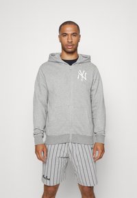 Sudadera gris con cremallera, con un bolsillo frontal y un logo blanco en el lado izquierdo del pecho, combinada con pantalones cortos a rayas grises y blancas con un detalle bordado.