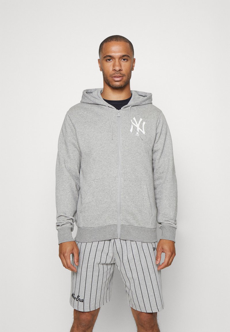 Sudadera gris con cremallera, con un bolsillo frontal y un logo blanco en el lado izquierdo del pecho, combinada con pantalones cortos a rayas grises y blancas con un detalle bordado.