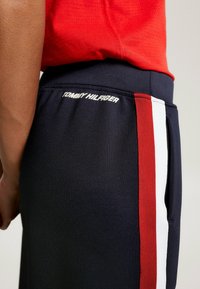 Marina blå träningsbyxor med en strukturerad tyg, elastisk midja och kontrasterande sidostråkar i rött och vitt. Logotypen visas på baksidan.