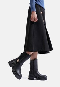 Stivali neri in pelle fino alla caviglia con suola spessa, zip frontale e accessori decorativi. Abbinati a una gonna nera a trapezio e un maglione a costine blu.