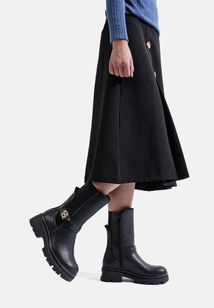 Stivali neri in pelle fino alla caviglia con suola spessa, zip frontale e accessori decorativi. Abbinati a una gonna nera a trapezio e un maglione a costine blu.