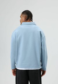 Hellblauer Pullover mit Kragen, lockerer Passform, langen Ärmeln und weicher, glatter Textur. Trägt ihn über einem weißen Hemd und dunkler Hose.