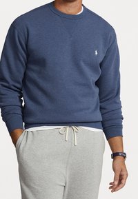 Marinblå sweatshirt i mjukt tyg, med rund halsringning, ribbade ärmslut och en liten logotyp på bröstet. Matchas med ljusgrå sweatpants.