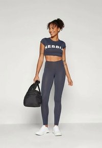 Ναυτικό crop top με κείμενο "NEBBIA", συνδυασμένο με ελαστικά σκούρα κολάν; το μοντέλο κρατάει μια μαύρη τσάντα γυμναστηρίου και φοράει λευκά αθλητικά παπούτσια.