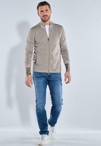 Beige Kapuzenjacke mit Stehkragen, kombiniert mit blauen Jeans. Weiße Sneakers setzen einen Kontrast. Verfügt über ein schlankes, tailliertes Design.