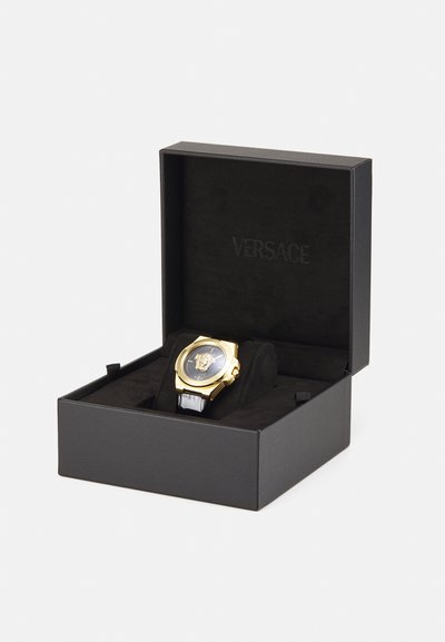 Versace laikrodis juodame dėkle. Jame yra auksinis korpusas, juodas ciferblatas su Medūzos logotipu ir juoda odinė apyrankė. Dėklo vidus išklotas aksomu.