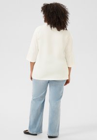 Top de manga larga color crema claro con un corte relajado, combinado con jeans rectos de color azul claro y bailarinas negras, visto desde atrás.
