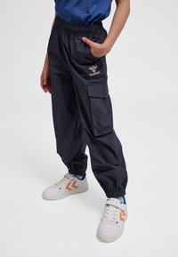 Copil purtând pantaloni cargo negri cu talie elastică, buzunar lateral și adidași albi cu detalii portocalii în formă de chevron, stând cu o mână în buzunar.