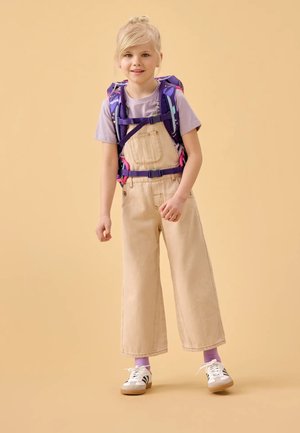Salopette beige in denim con gambe larghe, t-shirt viola chiaro e zaino con accenti viola. Scarpe da ginnastica bianche con strisce nere e calzini viola.